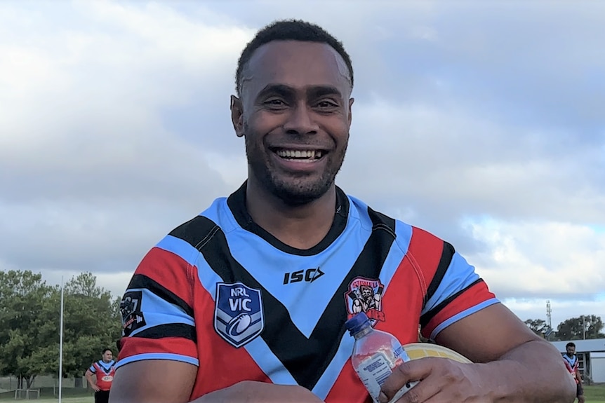 A man wearing a sky blue and red jersey holding a rugby ball and water bottle smiles