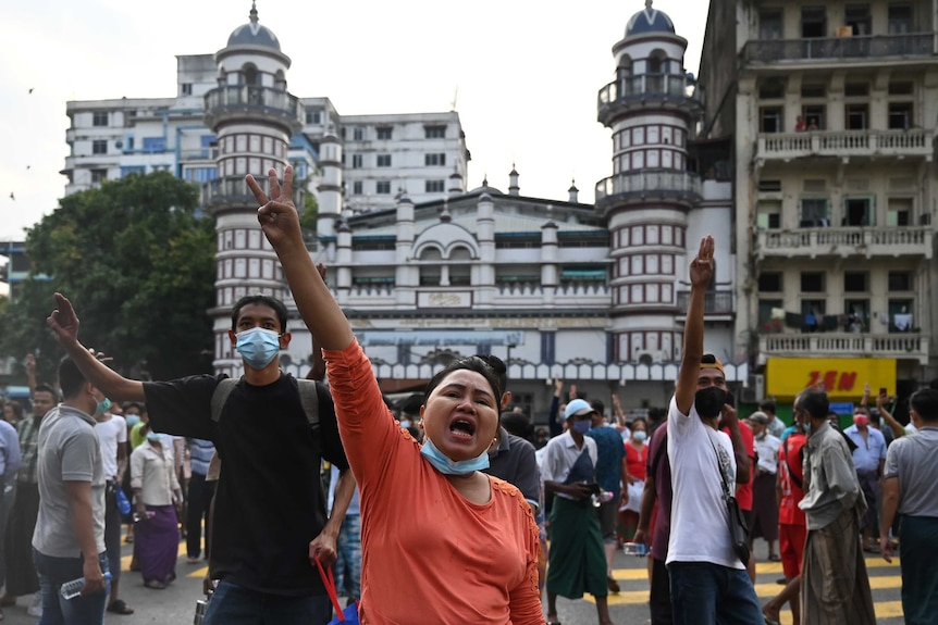 A woman and two men protest a military coup by raising three fingers on their right hands as they move among a large crowd.