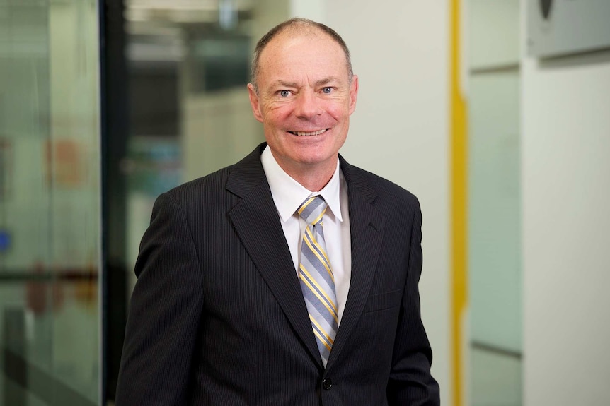 Victorian Coroner John Cain stands wearing a black suit smiling at the camera.