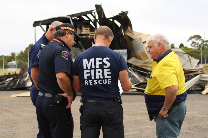 A policeman, CFS representative, MFS rep and a man in a high-vis shirt stand talking in front of the burned down building.