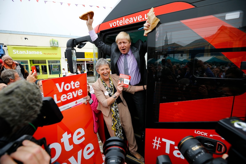 Boris Johnson holds up a pasty while standing behind a woman on a big red bus.