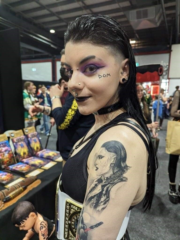 A woman with purple eyeshadow and black hair shows a black shoulder tattoo of wrestler Rhea Ripley.