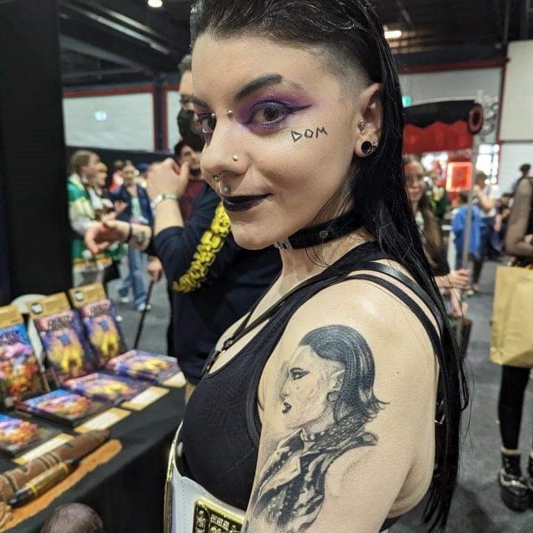 A woman with purple eyeshadow and black hair shows a black shoulder tattoo of wrestler Rhea Ripley.