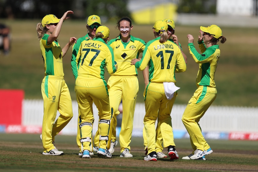 Megan Schutt smiles and is hugged by other women wearing yellow and green cricket kit