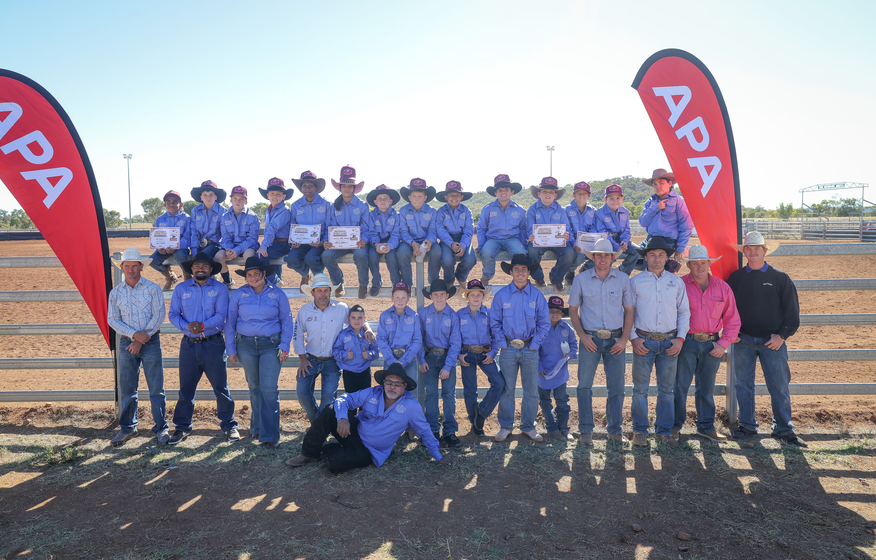 A large group of young riders and instructors pose together beside the arena after a junior rodeo clinic.