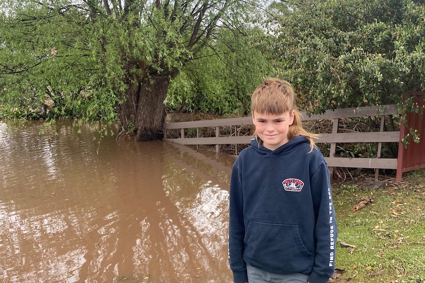 A smiling young lad with a spectacular mullet stands next to a flooded waterway.
