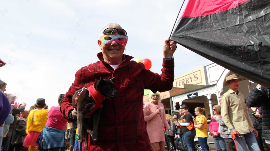 Laine marches in a LGBT parade in Broken Hill holding a small tent and dog and wearing a mask