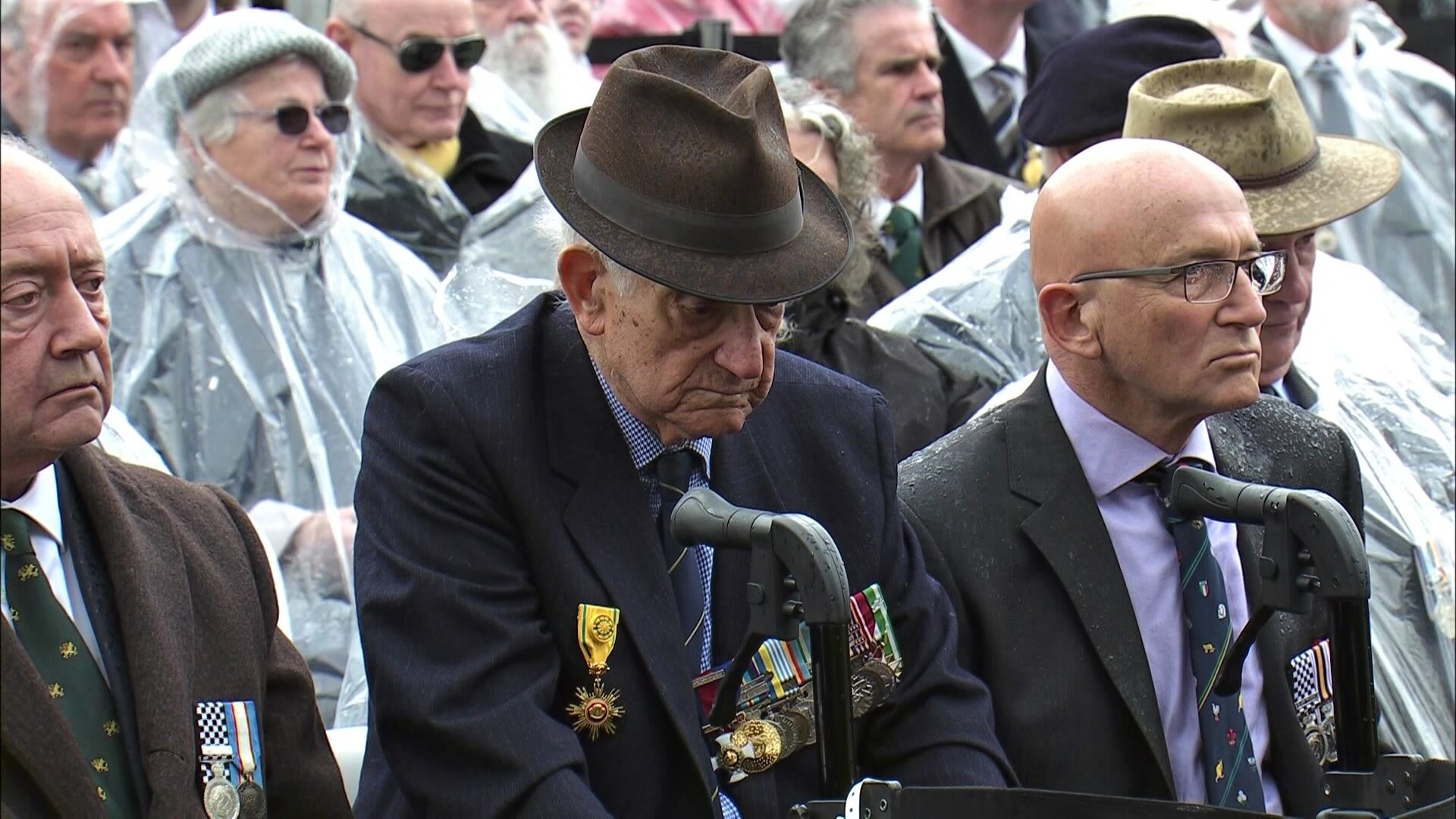 A group of men sit solemnly on chairs lined up at the service, some in uniform and wearing medals