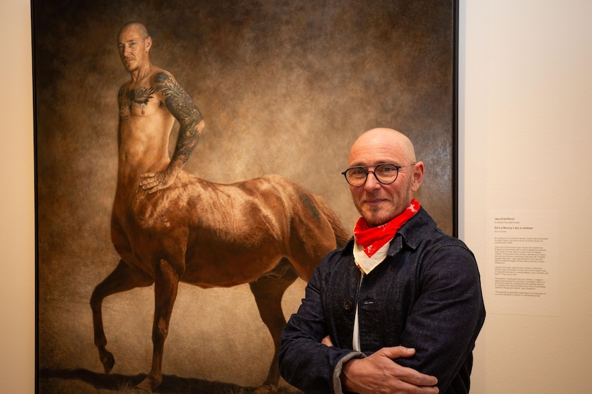 Ed Le Brocq, smiling with arms crossed across chest, with black rimmed glasses, stands before large portrait of him as centaur.