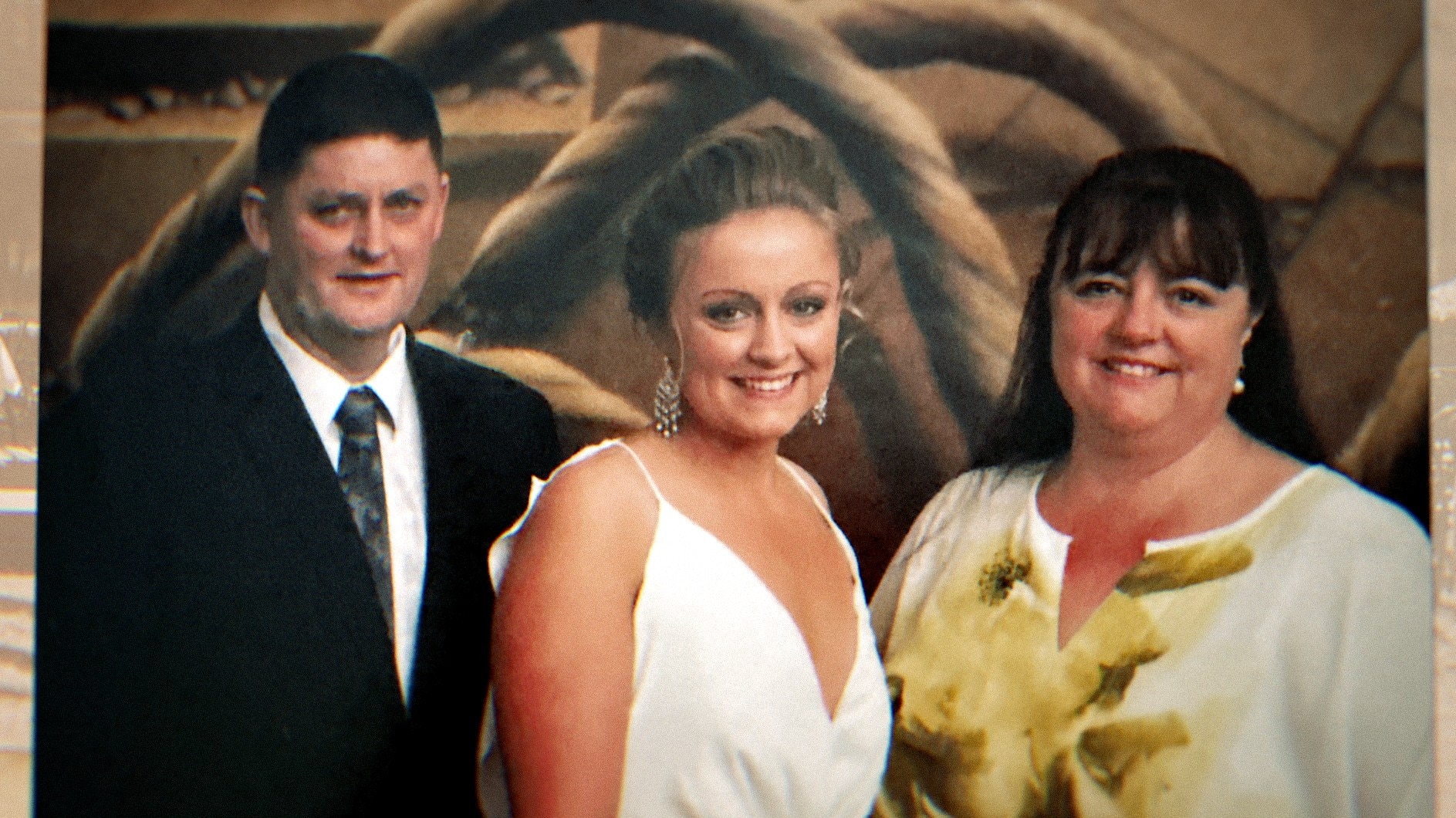A young woman wears a white dress, her blonde hair tied back. Her mother and father are either side and wear formal attire.