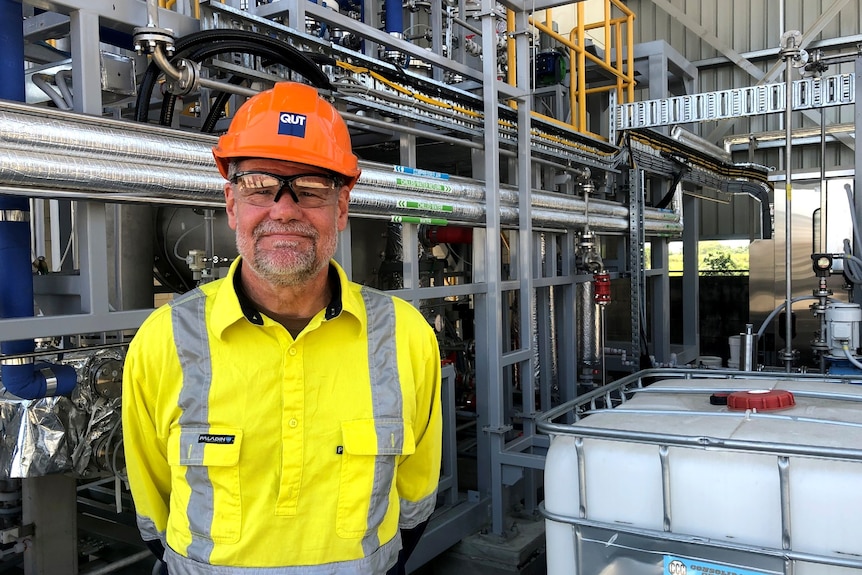 A man in hi-vis and an orange hard hat looks at the camera in front of grey pipes. 