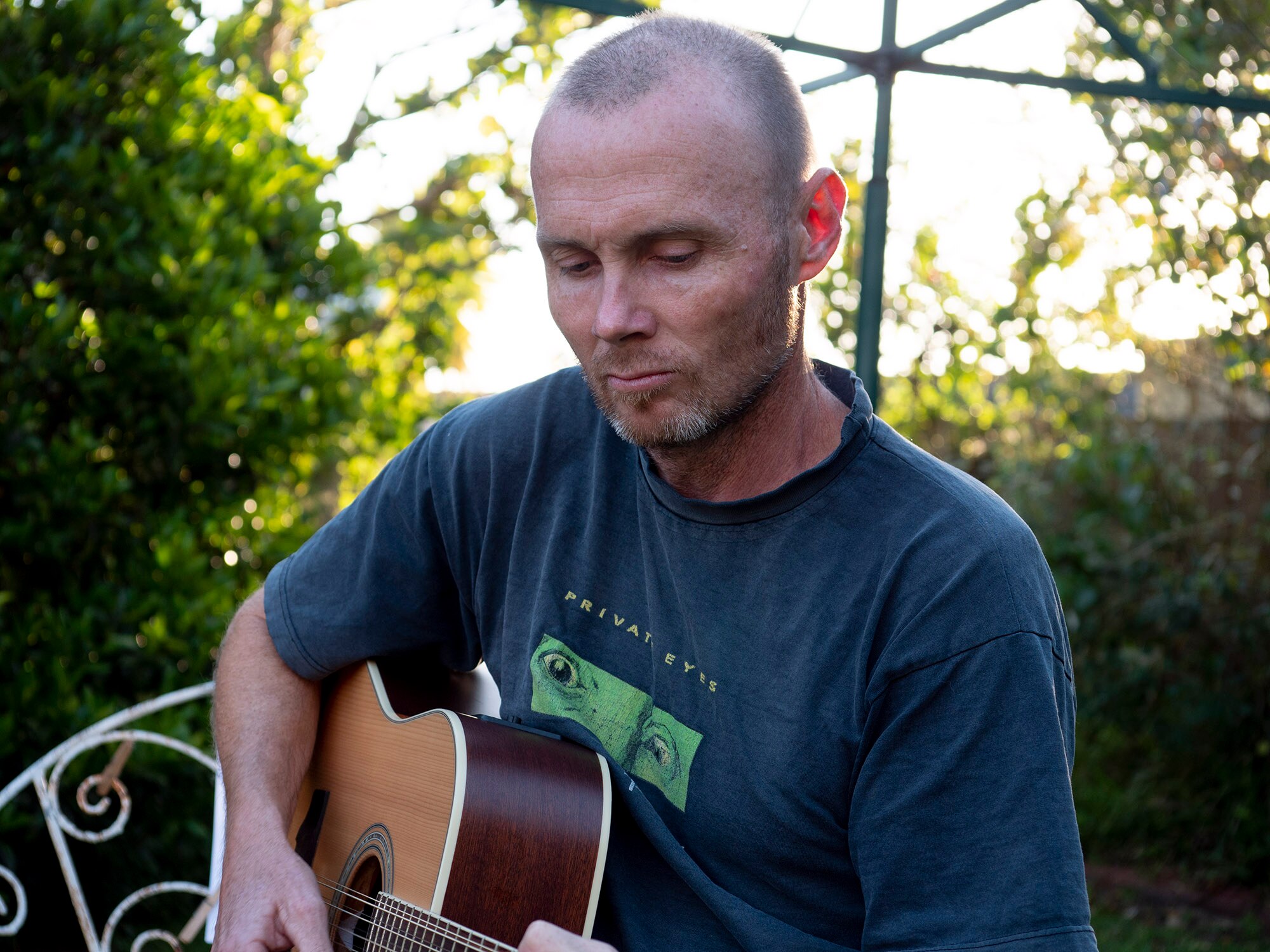 A male in a dark t-shirt playing a guitar in the backyard.