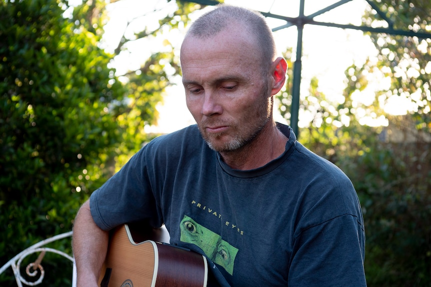 A male in a dark t-shirt playing a guitar in the backyard.