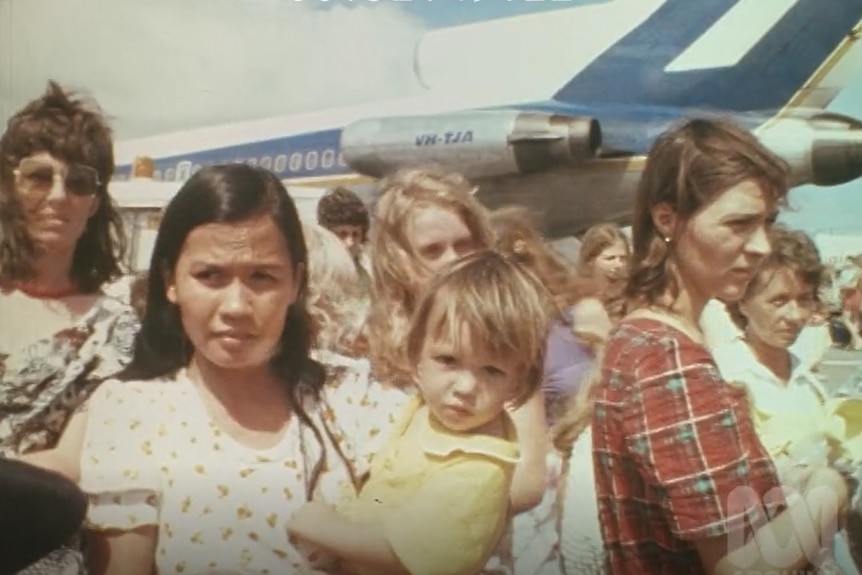 Woman holds child in a crowd with a plane behind.