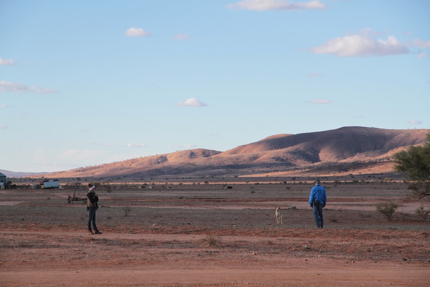 Actors stand on an outback landscape with hills in the background.