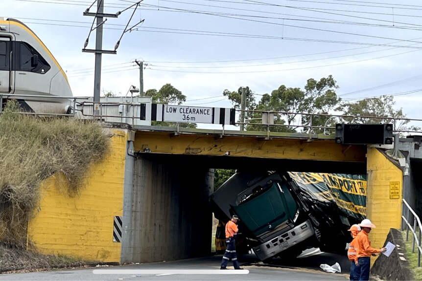 A truck stuck in a rail underpass, a train visible on the rail line above