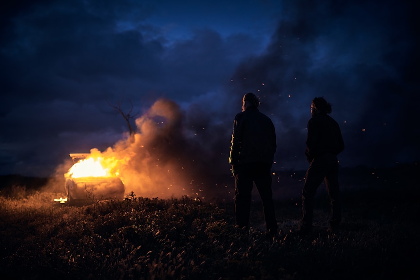A big log burns in the night as two middle-aged white men watch it in an open field
