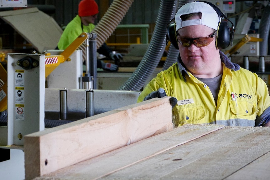 A man in high-vis wear, white cap and safety glasses saws a piece of wood