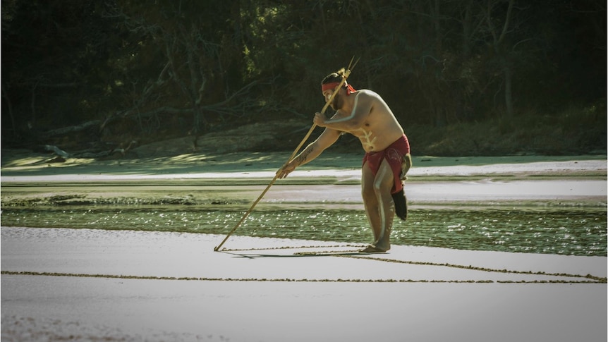 Indigenous man drawing a lined picture in the beach sand, in front on green weed and trees in background.