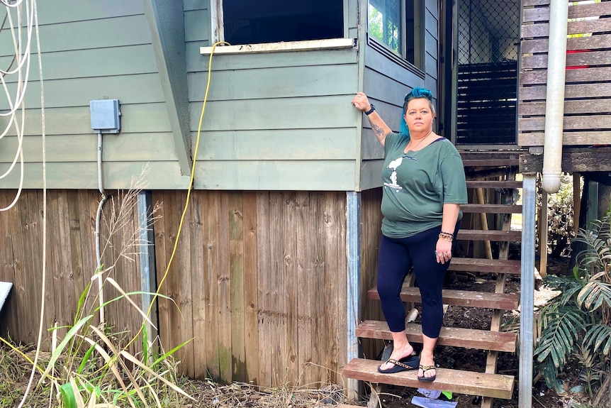 Terri wearing green top, black pants standing on steps holding her right hand up to a level on the house where the water reached