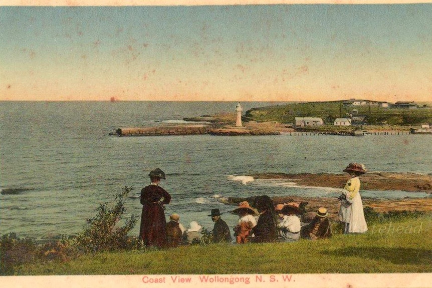 A vintage postcard depicts a view of the harbour with people on a green hillside looking across to the lighthouse and headland