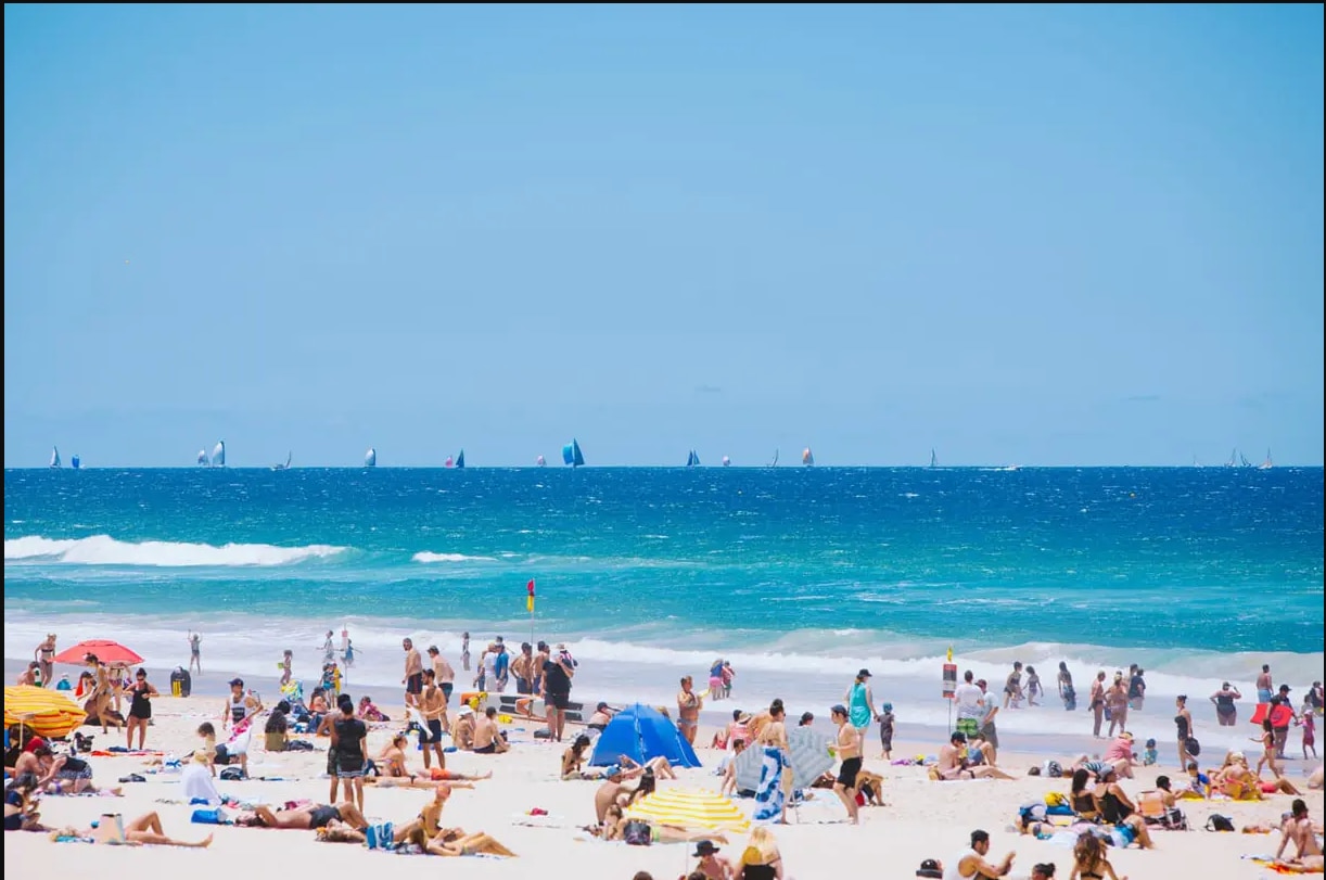 Dozens of people relaxing and playing on a beach with sail boats on the horizon.