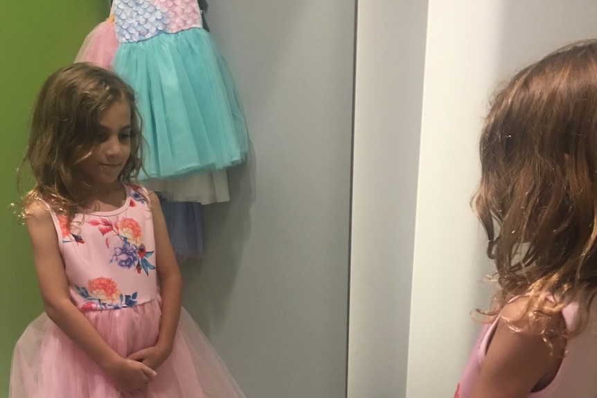 a young girl stands in a change room wearing a pink dress and looking at herself in the mirror