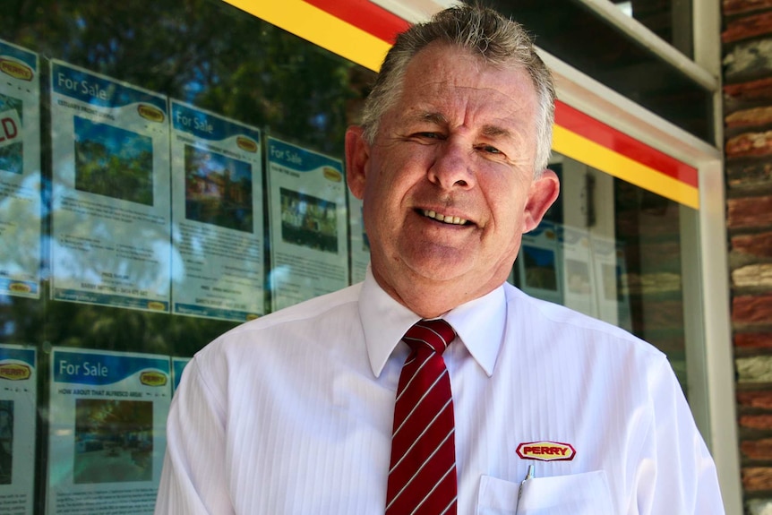 a head and shoulders shot of a man in a white shirt and red tie standing outside a real estate agency.