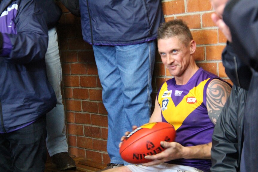 A man with short blonde hair and a tattooed shoulder wears an AFL guernsey and holds the ball in the dugout. 
