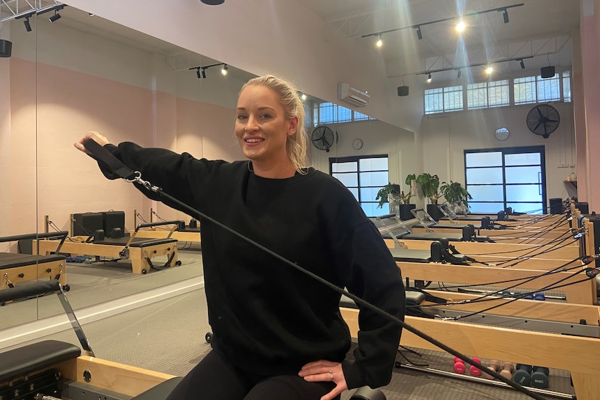 A blonde woman in black clothes works an exercise band in a Pilates studio.