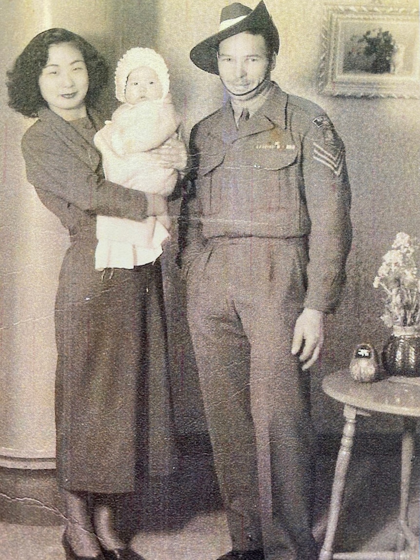 A black and white photo of a Japanese woman holding her baby standing next to an Australian male soldier in uniform