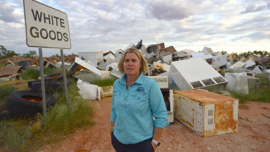 A lady with blonde hair in a blue shirt standing next to a large pile of white goods at a dump and a sign saying white goods