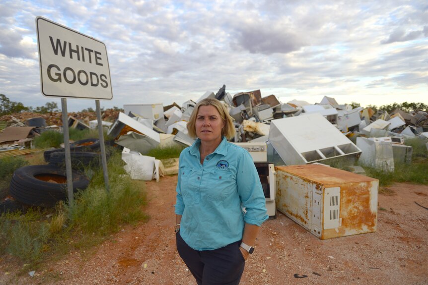 A lady with blonde hair in a blue shirt standing next to a large pile of white goods at a dump and a sign saying white goods