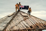 Expeditioners hoist the Australian flag over an old wooden hut.