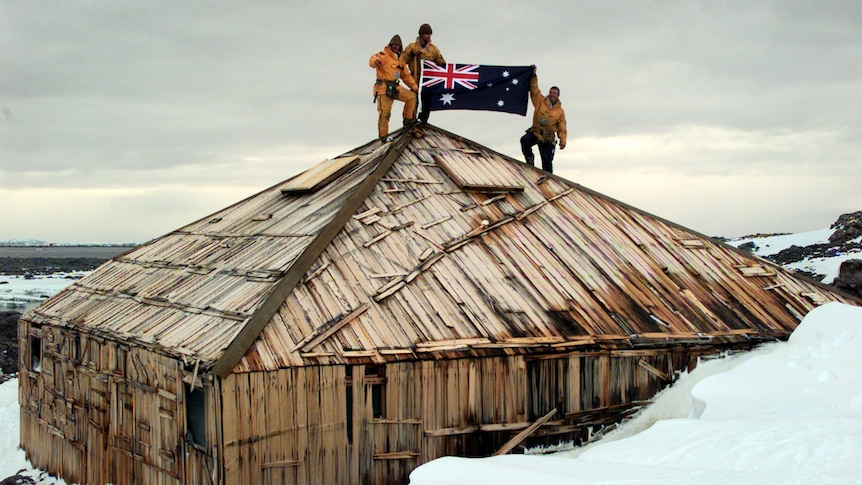 Expeditioners hoist the Australian flag over an old wooden hut.