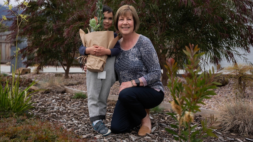 A woman kneels with her grandson on a nature strip covered by bark chips and plants.