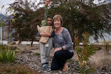 A woman kneels with her grandson on a nature strip covered by bark chips and plants.