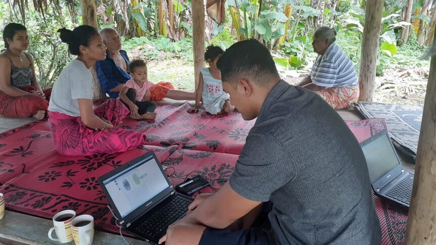 A young man sits crossed legged with a laptop in a traditional Samoan house surrounded by a multi-generational family.