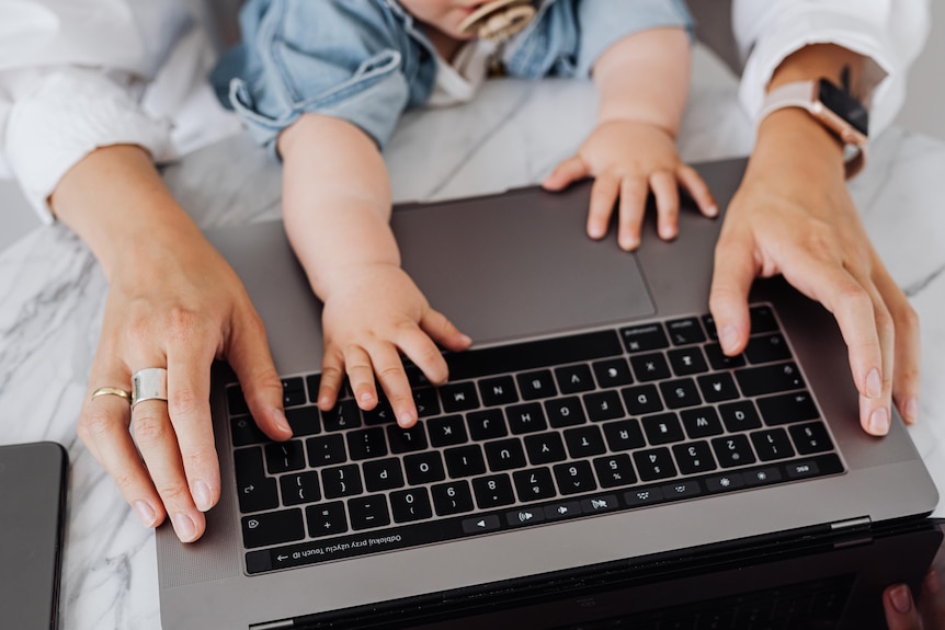 A woman's hands on a black laptop keyboard, with two chubby baby hands reaching for the keys in between. 
