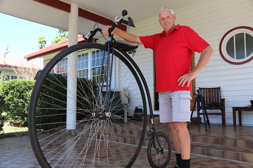 A man in a red shirt resting his hand on the handlebars of a large penny farthing