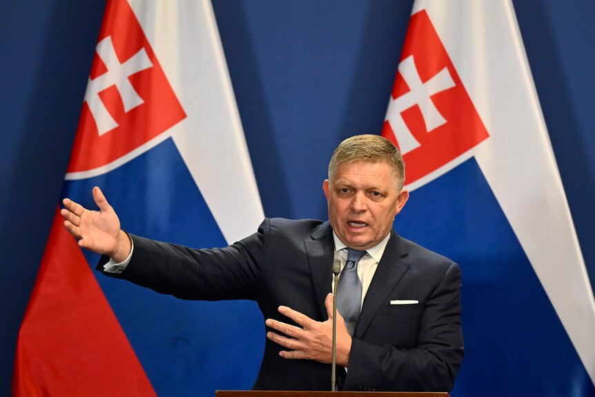 A middle-aged man in a suit gestures as he speaks in front of two Slovakian flags.