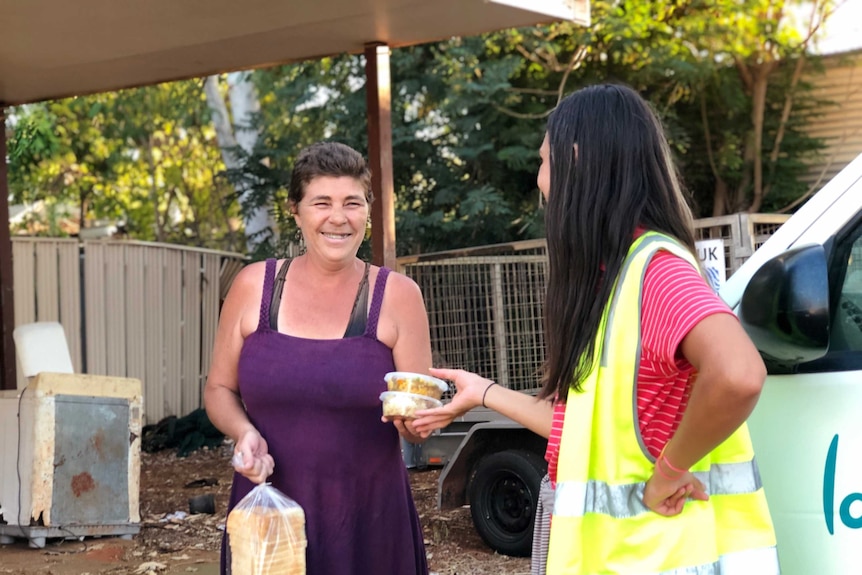 A woman in a fluro vest hands over containers of food to a woman in a purple top, in a driveway in Broome