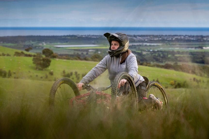 A woman on a three-wheeled bike wearing a helmet and smiling. Scenic hills are in the background.