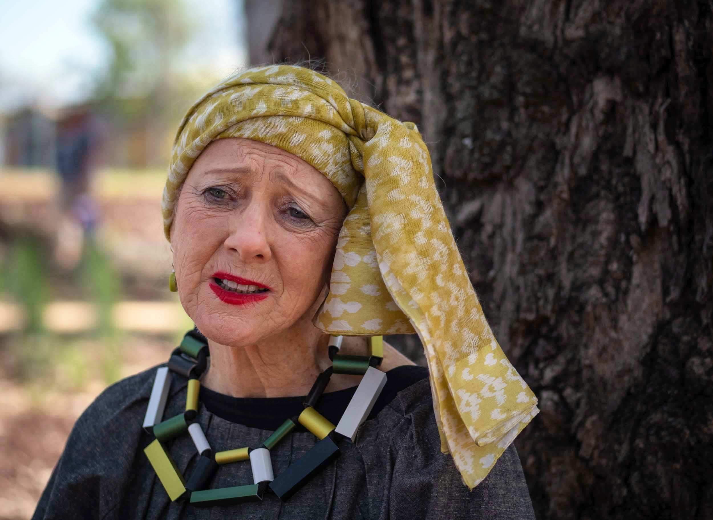 a woman in a colourful headscarf and blocky necklace has a concerned expression on her face.
