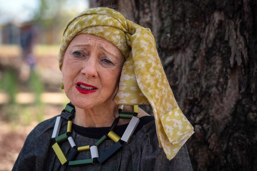 a woman in a colourful headscarf and blocky necklace has a concerned expression on her face.