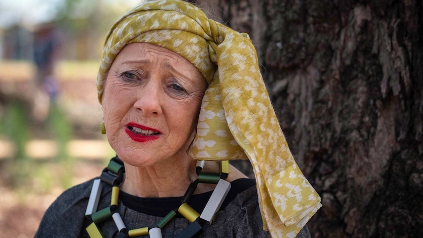 a woman in a colourful headscarf and blocky necklace has a concerned expression on her face.