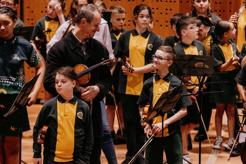 Richard Tognetti, a white man in his 60s, holds a violin beside Lachlan Martin, an 11-year-old white boy, also holding a violin.