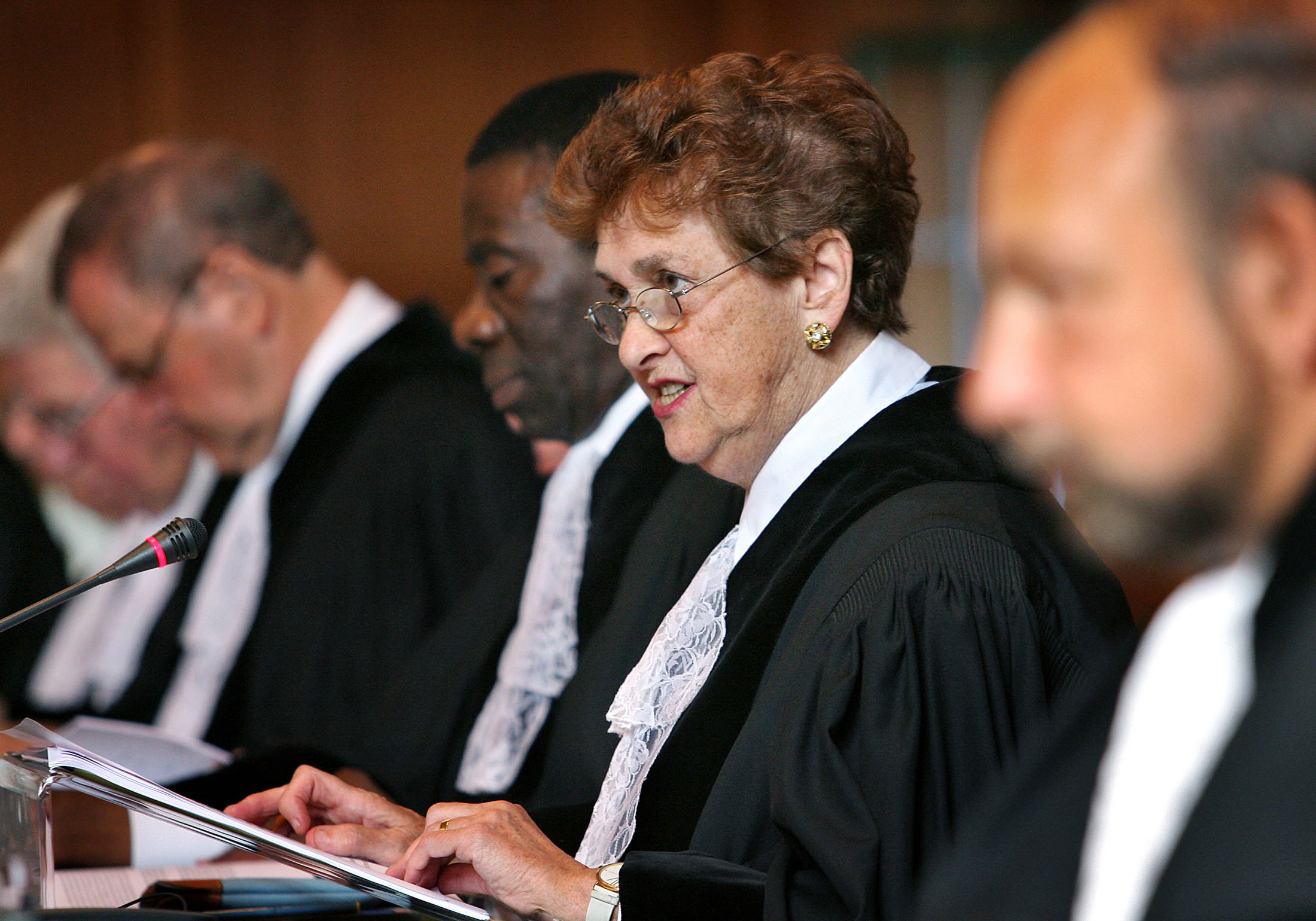 Rosalyn Higgins, with short brown hair, black cloak and white neck-tie speaks while blurred figures sit to the side of her.