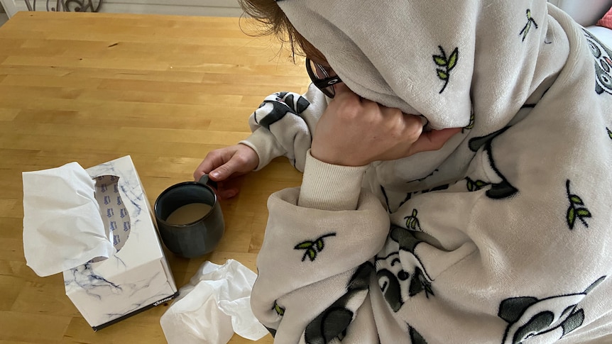 Person leaning over a table wearing a hooded gown, with a box of tissues and a mug on the table.
