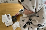 Person leaning over a table wearing a hooded gown, with a box of tissues and a mug on the table.